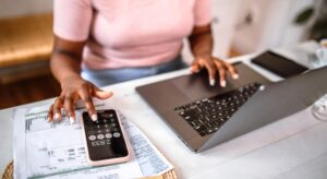 Close up of woman using phone as a calculator while typing on her laptop